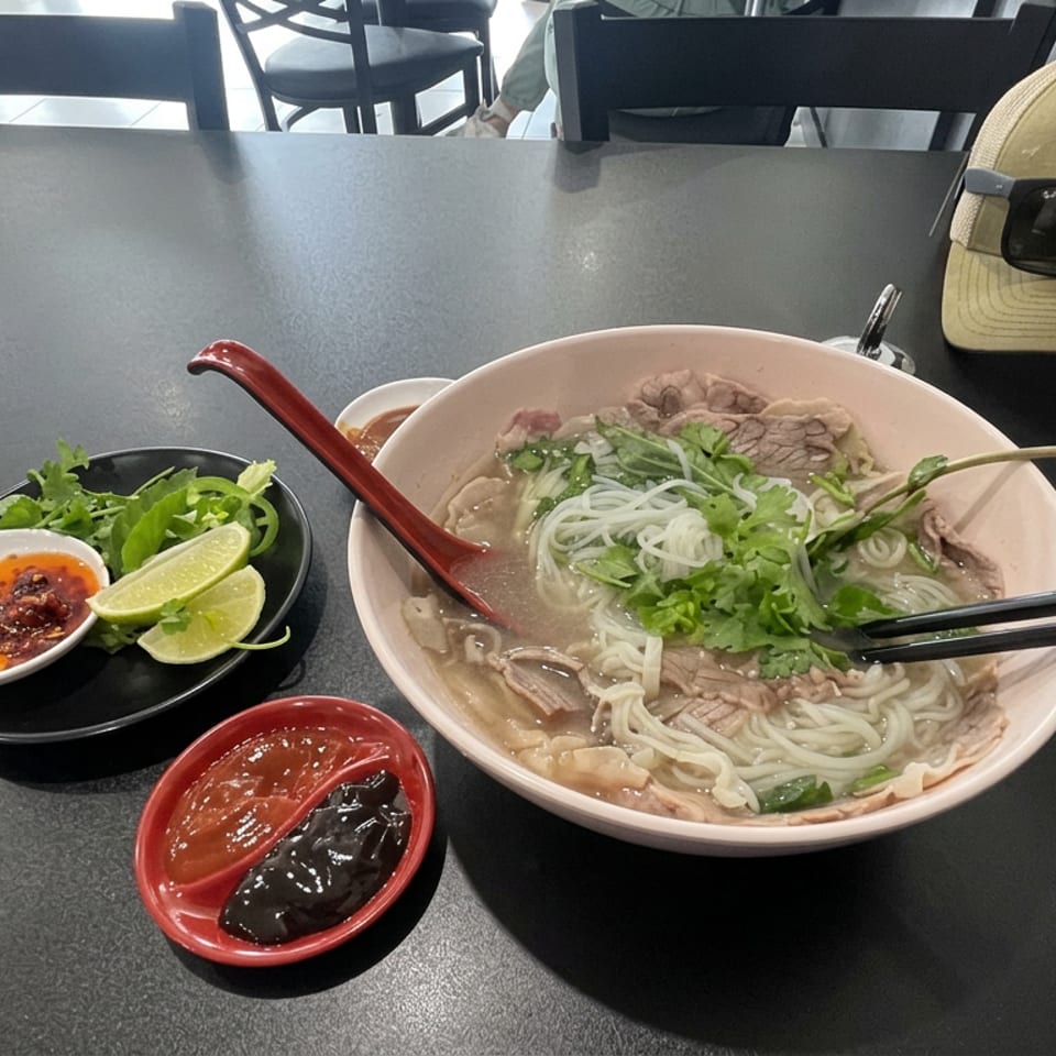 Vietnamese pho with sliced beef, rice noodles, herbs, lime, chili, and sauces served on a restaurant table.