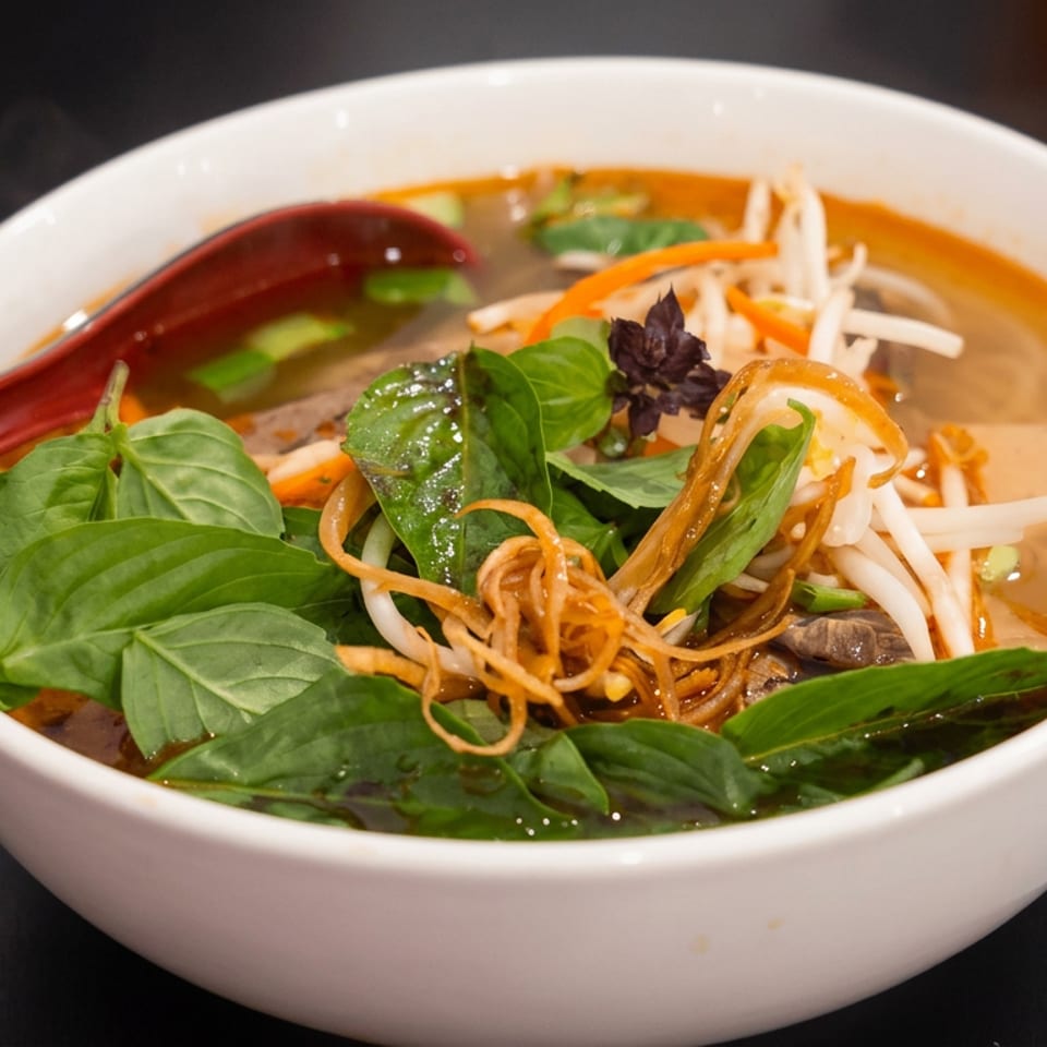 Bowl of steaming Vietnamese noodle soup topped with basil, bean sprouts, carrots, mushrooms, and a red spoon.